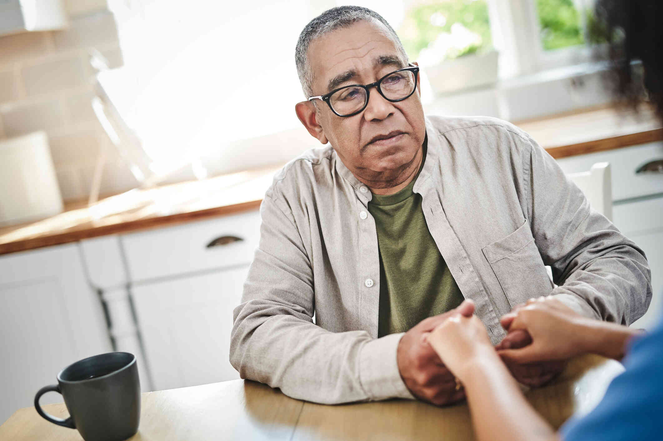 An elderly man in glasses sits at atable in his home and holds the hands of the relative sitting across from him with a worried expression.