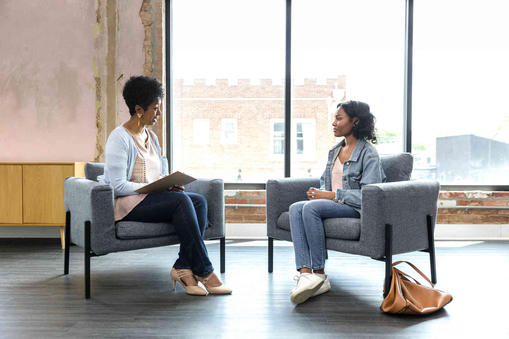 A woman in a jean jacket sits in a chair across from her female therapist during a therapy session.