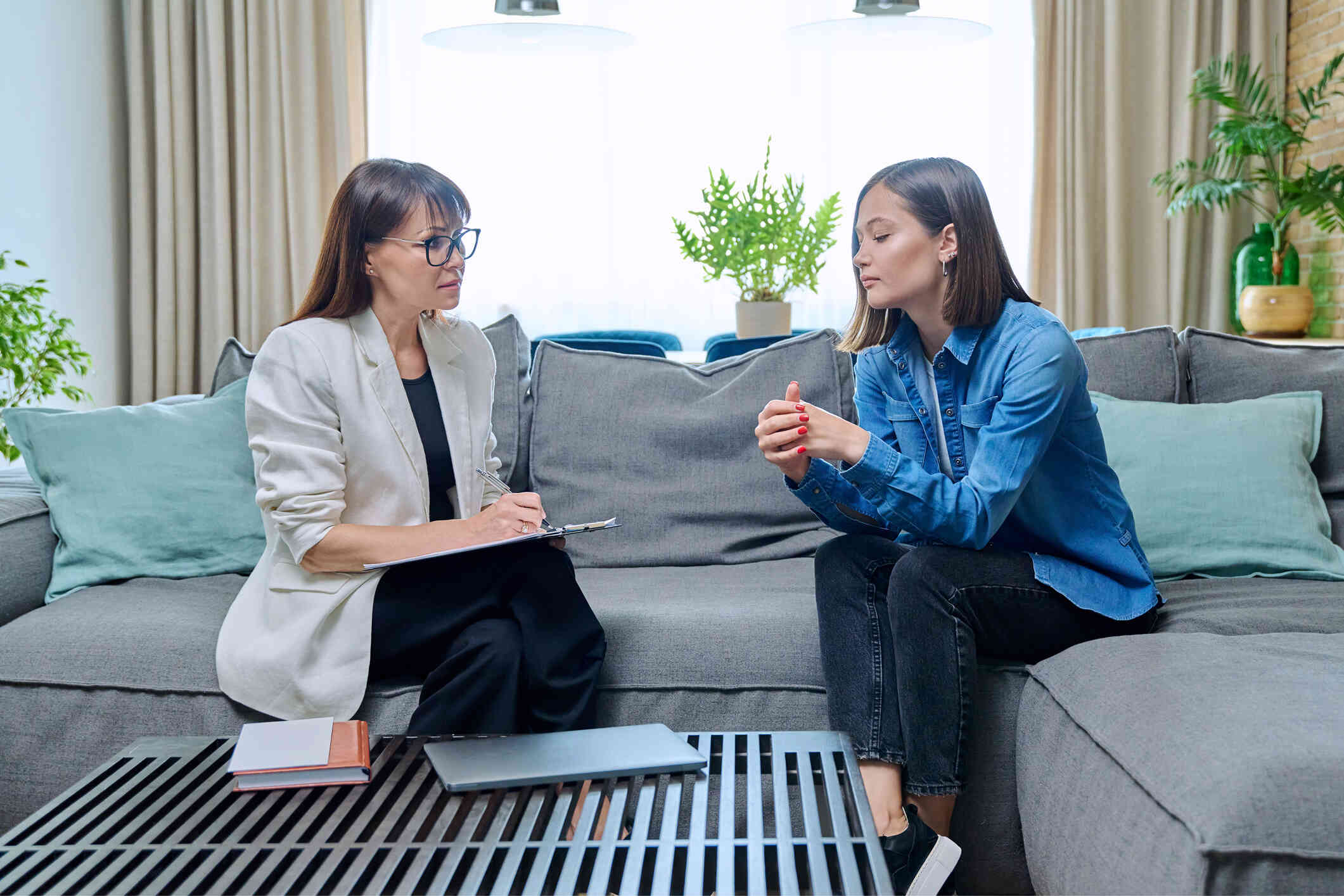 A woman in a jean shirt leans forward and talks to her female therapist while sitting on the couch next to her.