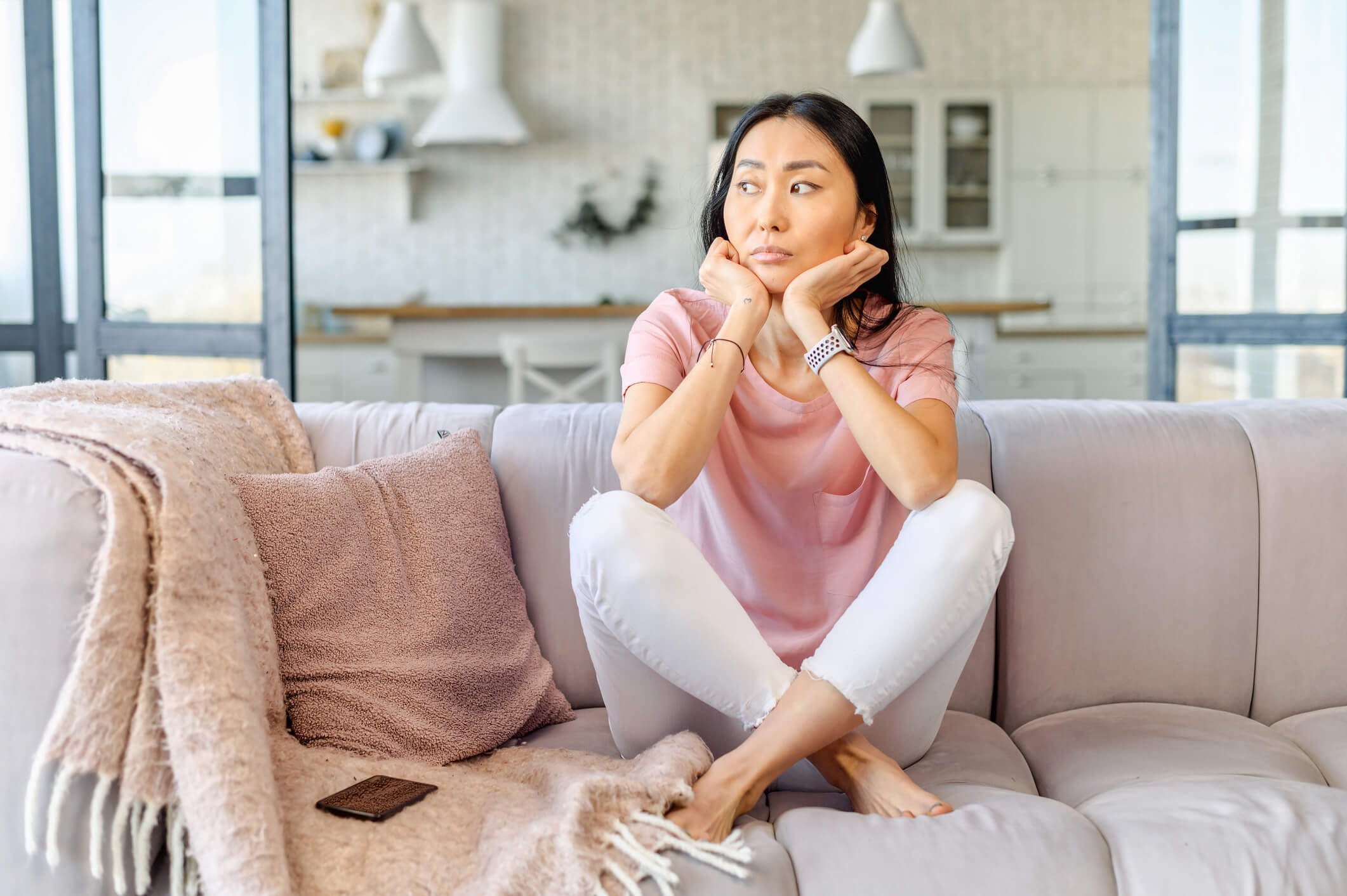 A woman is sitting on a couch with her feet up; she has her hands on her face, is looking away, and has a sad expression.